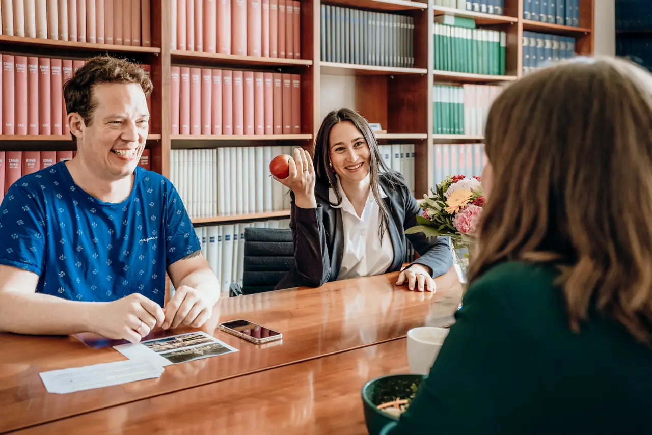 Drei Mitarbeitende von RIFA lachen am Tisch im Besprechungsraum vor Regalen mit Fachliteratur, eine Kollegin hält einen Apfel