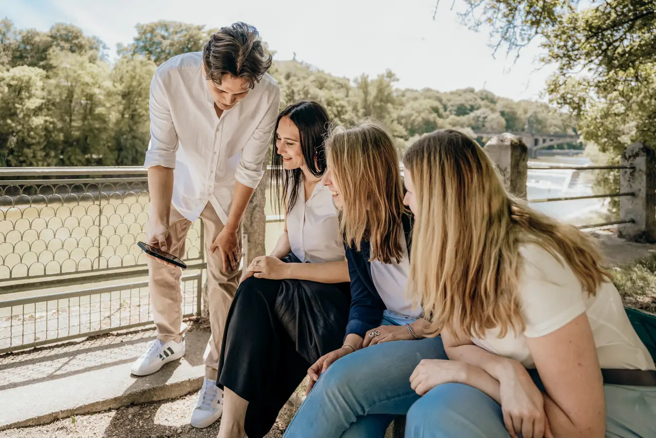 Ein Mitarbeiter zeigt drei Kolleginnen auf einer Bank an der Isar etwas auf dem Smartphone, alle lachen