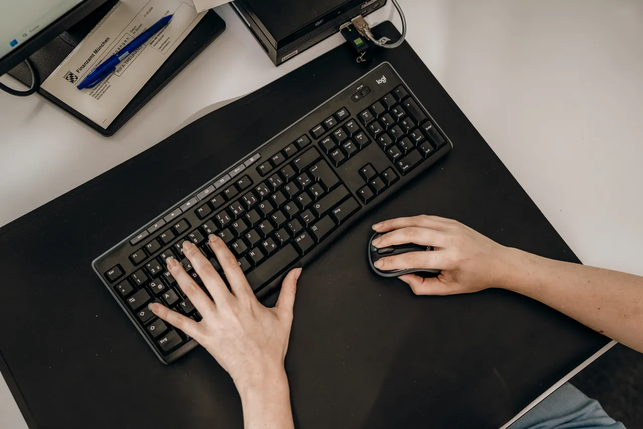 Hände an Tastatur am Schreibtisch, daneben Unterlagen und Stift